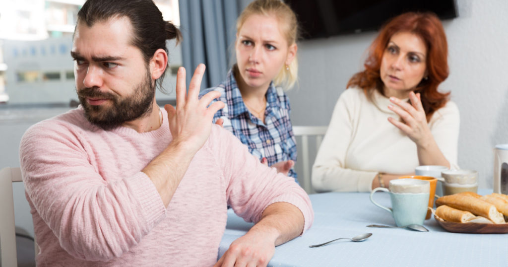 Upset young man sitting at table after quarrel and girl with mother on background at home