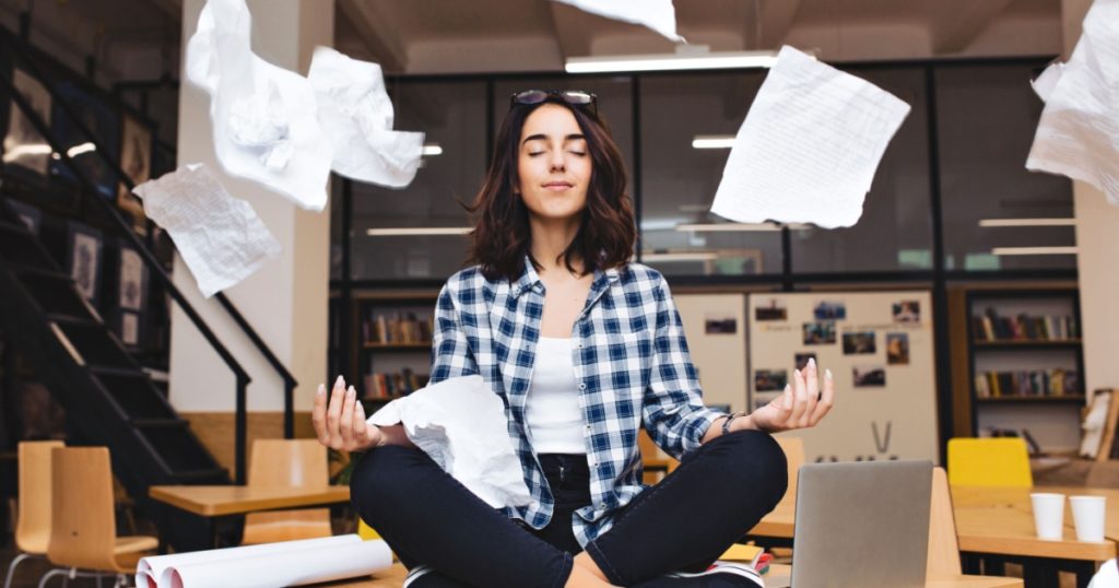 Young pretty joyful brunette woman meditating on table surround work stuff and flying papers. Cheerful mood, taking a break, working, studying, relaxation, true emotions
