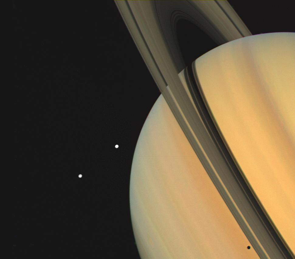 Saturn and two of its moons, Tethys (above) and Dione, were photographed by Voyager 1 on November 3, 1980, from 13 million kilometers (8 million miles). The shadows of Saturn's three bright rings and Tethys are cast onto the cloud tops. The limb of the planet can be seen easily through the 3,500-kilometer-wide (2,170 mile) Cassini Division, which separates ring A from ring B. The view through the much narrower Encke Division, near the outer edge of ring A is less clear. Beyond the Encke Division (at left) is the faintest of Saturn's three bright rings, the C-ring or crepe ring, barely visible against the planet.