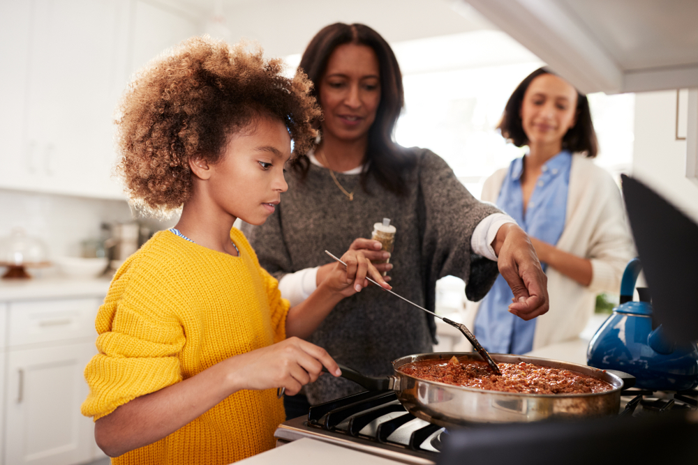 Pre-teen African American girl standing at the hob in the kitchen preparing food with her grandmother and mother, close up, selective focus