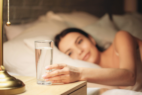 Beautiful young woman taking glass of water from bedside table in evening