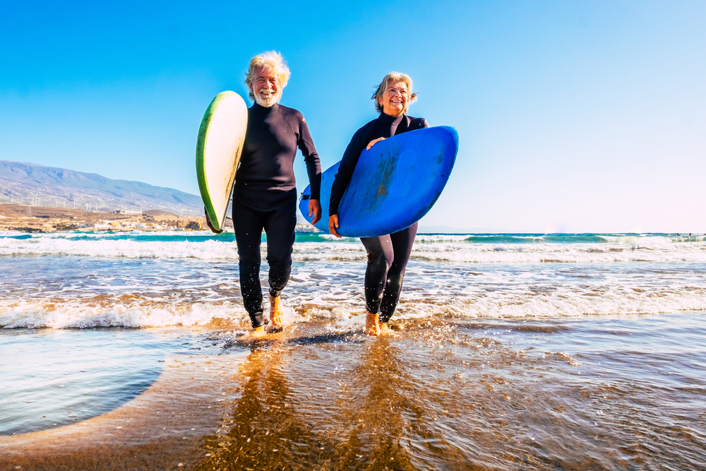 two old and mature people having fun and enjoying their vacations outdoors at the beach wearing wetsuits and holding a surfboard to go surfing in the water with waves - active senior smiling