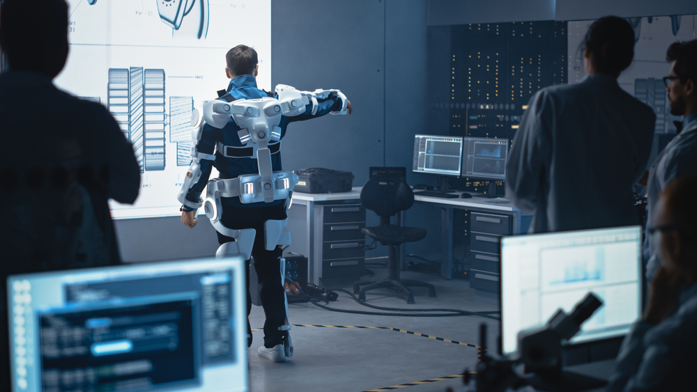 In Development Laboratory: Engineers and Scientists Look at Robotics Exoskeleton Prototype Presentation with Person Testing it, Moving and Performing. Designing Wearable Exosuit to Help People