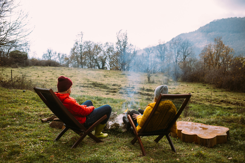Couple in warm puffy jackets sit on sun chairs in front of cozy campfire at campground or country house. Off grid living in nature. Millennial weekend getaway to camp overnight location
