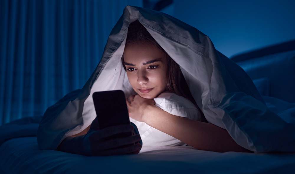 Young female covered with blanket lying in bed and browsing internet on mobile phone before sleep at late night