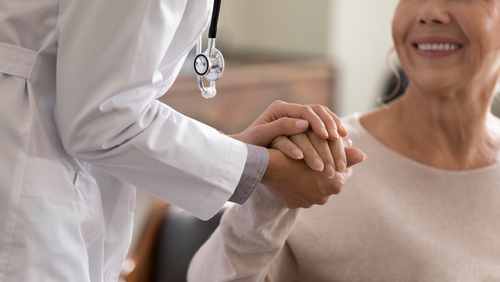 Doctor giving hope. Close up shot of young female physician leaning forward to smiling elderly lady patient holding her hand in palms. Woman caretaker in white coat supporting encouraging old person
