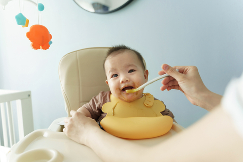 Happy Smiling Asian Little cute baby boy wearing yellow bib enjoying eating porridge or pureed food while sitting on high chair at home. Cheerful Lovely Mom feeding her son with baby spoon. BeH3althy
