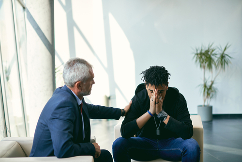 Unhappy black adolescent talking about his problems with mental health professional during therapy at counselling center.