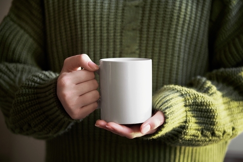 female hand holding white mug with blank copy space for your advertising text message or promotional content, sweet coffee or tea. Girl in green sweater holding white porcelain mug mock up
