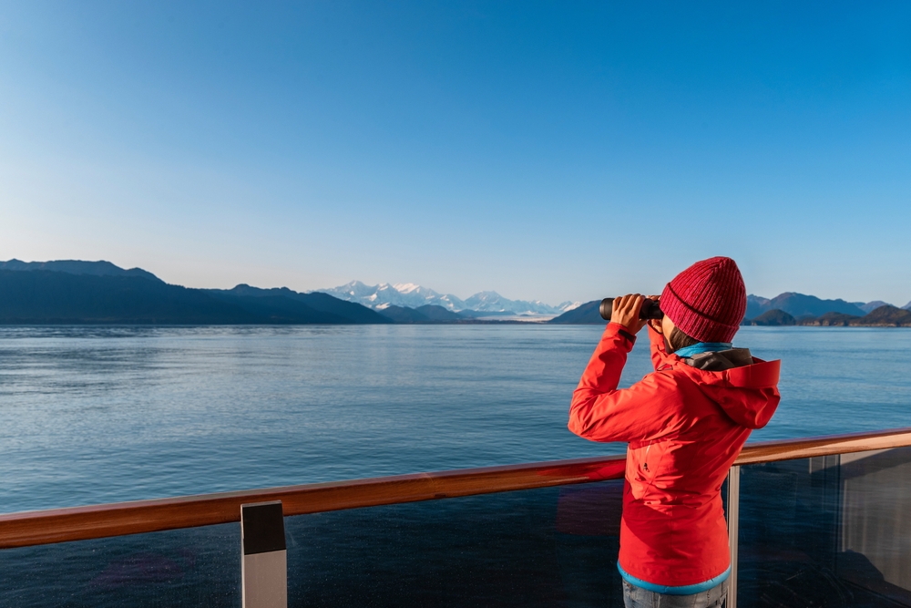 Vacation adventure. Alaska Glacier Bay cruise ship passenger looking at Alaskan mountains with binoculars exploring Glacier Bay National Park, USA. Woman on travel Inside Passage enjoying view