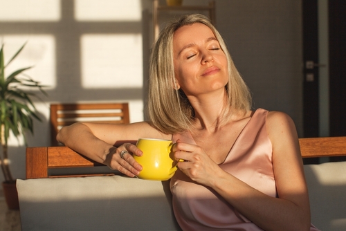 Attractive happy middle aged woman is sitting on sofa in living room. Smiling adult lady enjoys drinking coffee or tea sitting on couch at home