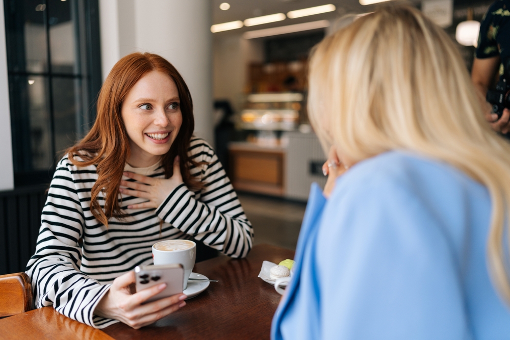 Side view of two happy beautiful women friends sitting in cafe indoors, looking at phone and talking, laughing. Cheerful pretty females girlfriends using social media on smartphone in coffee shop.