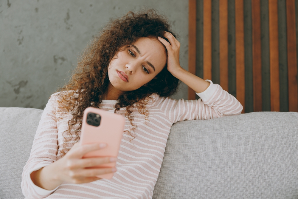 Close up young dissatisfied sad woman wear casual clothes sits on grey sofa couch hold in hand use mobile cell phone stay at home hotel flat spend free spare time in living room indoor. Lounge concept