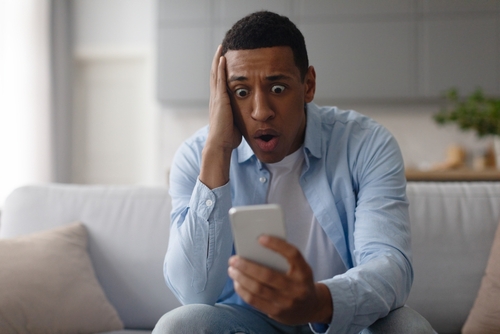 Shocked black man dealing with cellphone issue, looking at phone, reading unpleasant news or message, sitting on sofa at home and touching head