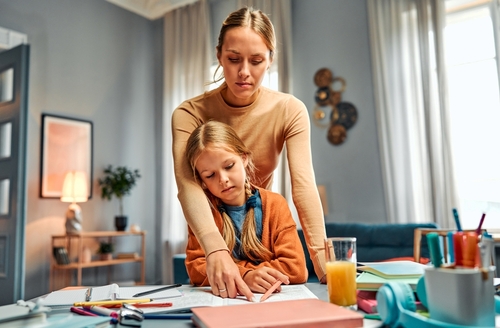 Concept of strict upbringing. Demanding mom hanging over kid from behind and pointing at mistake done in notebook. Serious child learning being attentive and responsible while doing home tasks.
