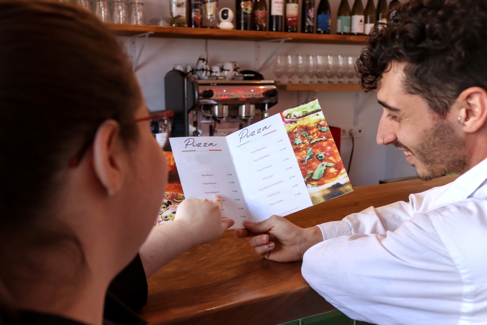 Person smiling choosing food from a menu receives recommendations from the waitress, seen from behind, in a pizza restaurant