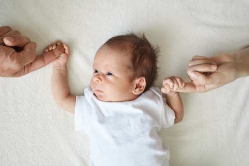 Baby holding parents hands. Father and mother hold newborn kid cute infant child in white bodysuit lying on white sheet on bed
