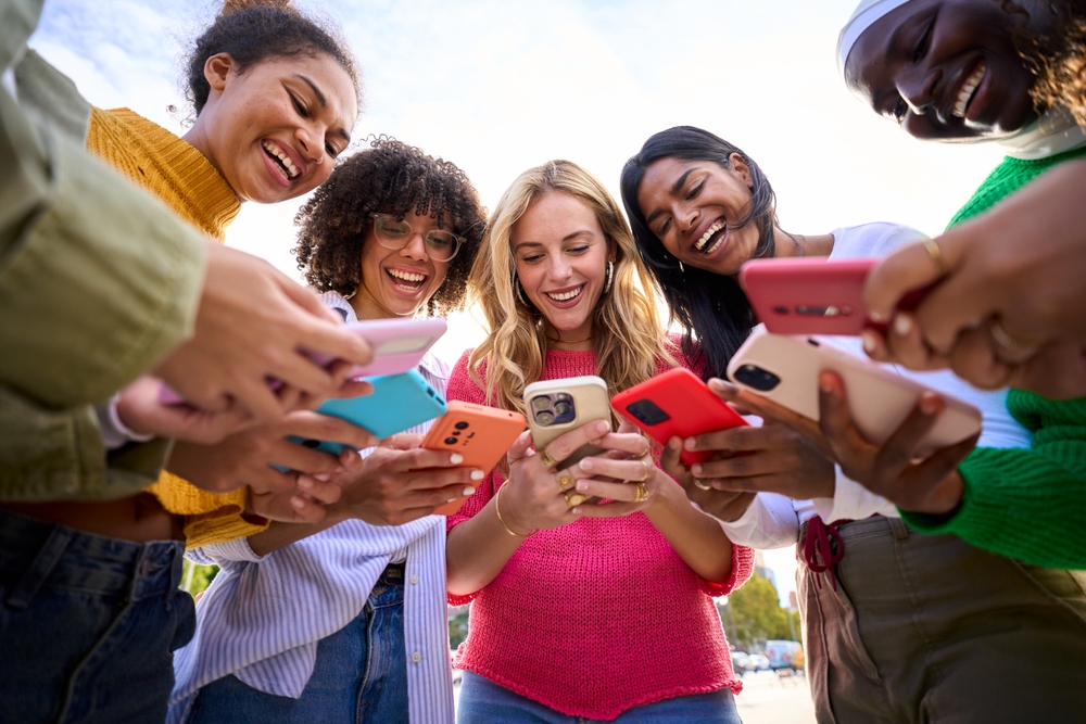 Group of international happy generation z only women laughing gathered in circle looking at cell phones outdoors. Cheerful young females using mobiles with funny expression. Addiction to technology