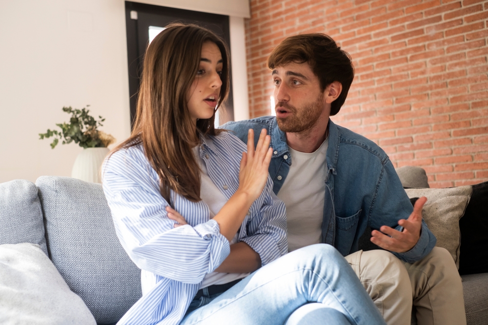Angry man shouting on phone at home expressing frustration and stress. Young adult with glasses having heated conversation displaying intense emotions in modern living room setting.