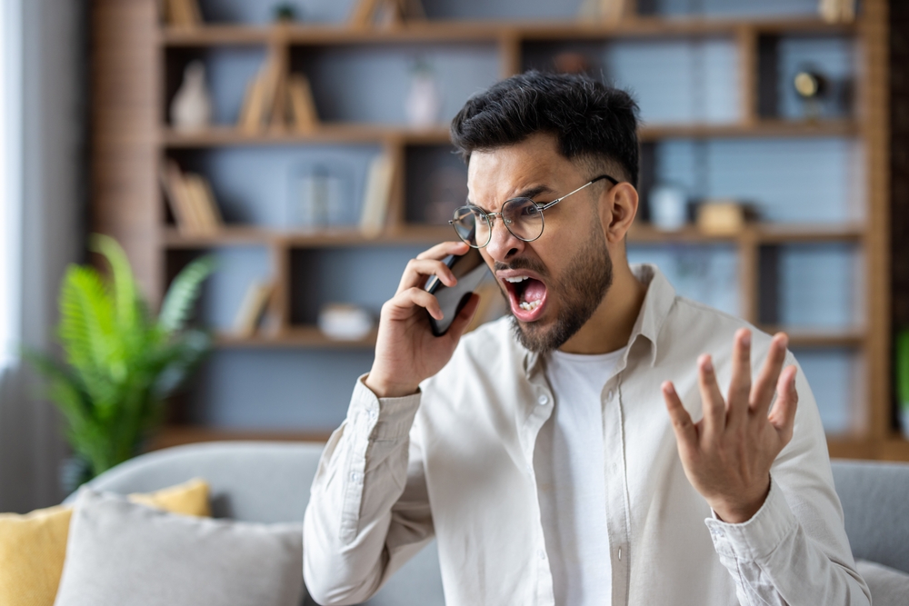 Angry man shouting on phone at home expressing frustration and stress. Young adult with glasses having heated conversation displaying intense emotions in modern living room setting.