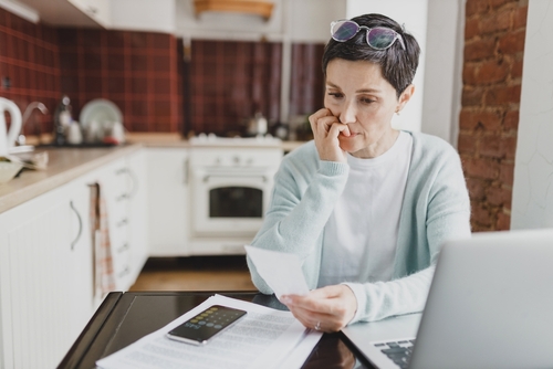 Selective focus on worried anxious female of 60s biting nails holding utility bill in hands sitting at kitchen table, trying to calculate expenditures, using laptop to pay and smartphone calculator