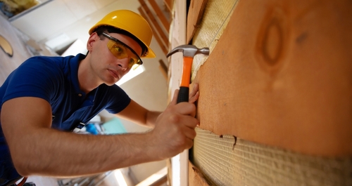 A construction worker wearing a yellow hard hat and protective glasses uses a hammer to secure insulation material in place. The unfinished attic space suggests ongoing renovation or building work.