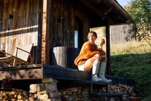 A woman in an orange sweater and boots sits on a wooden porch with a warm drink, enjoying the fresh morning air. Sunlight gently illuminates the cozy scene