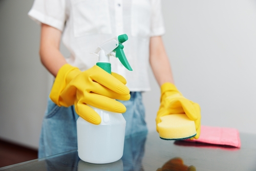 Woman in yellow rubber gloves holds cleaning solution and sponge on table with cloth