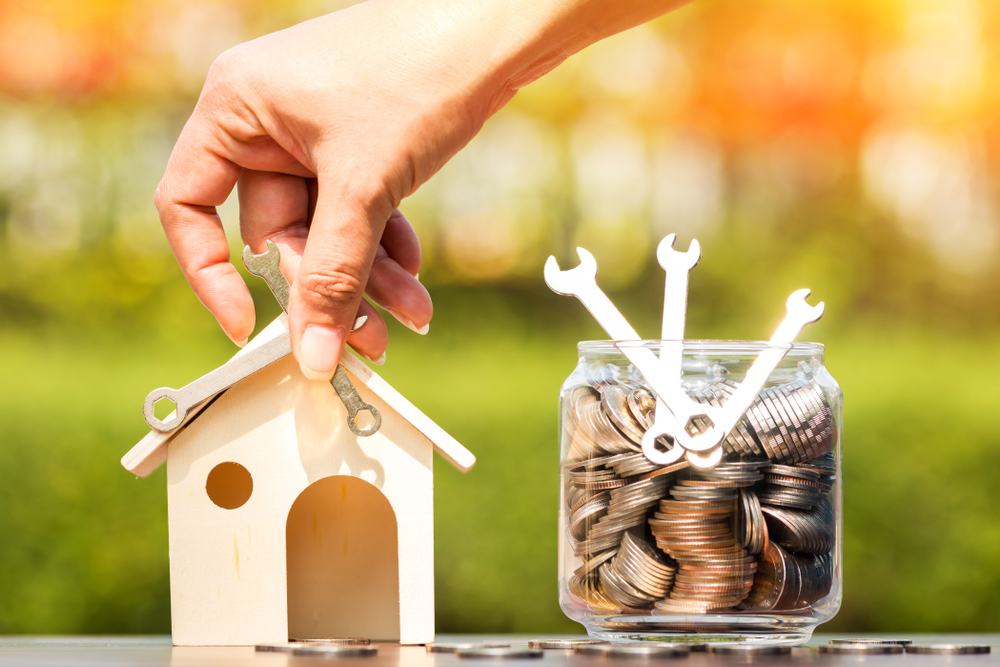 Women hand are repairing wooden house and a bottle bank with money and a wrench put on the wooden in the public park, saving money and loan for construction real estate and home concept.
