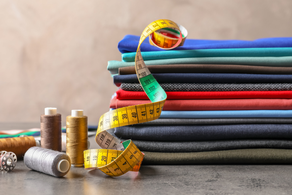 Bobbins with threads, measuring tape and stack of colorful fabrics on table. Tailoring accessories