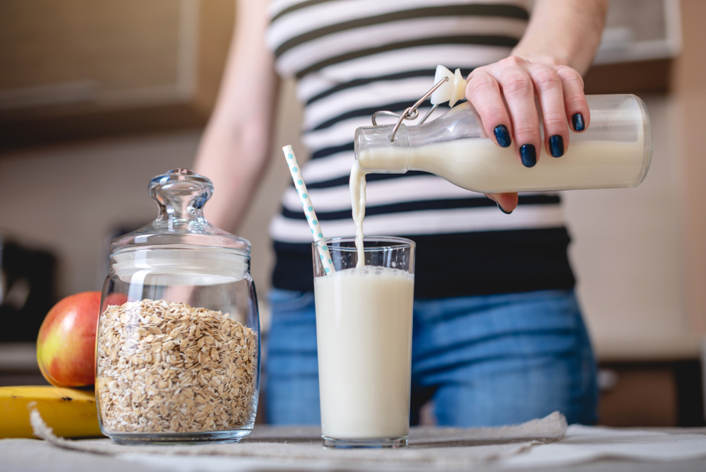 A woman pours organic oat milk from a bottle into a glass on a table in the kitchen. Diet healthy vegetarian product