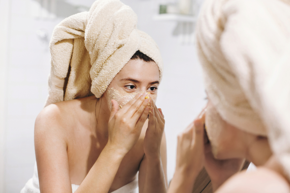Young happy woman in towel making facial massage with organic face scrub and looking at mirror in stylish bathroom. Girl applying scrub cream, peeling and cleaning skin. Skin Care