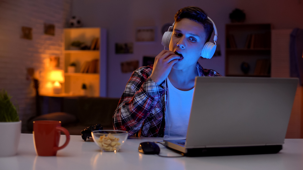 Excited teenage boy playing computer game and eating snack, leisure activity