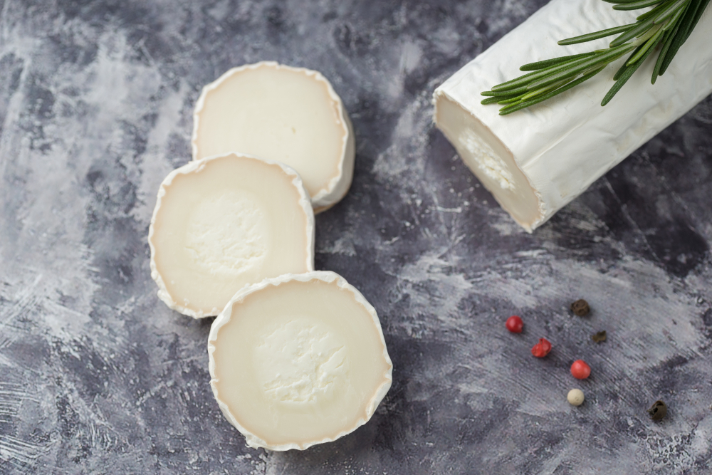 Delicious goat cheese slices and piece with fresh rosemary leaves on stone background. Luxurious culinary cheese still life.