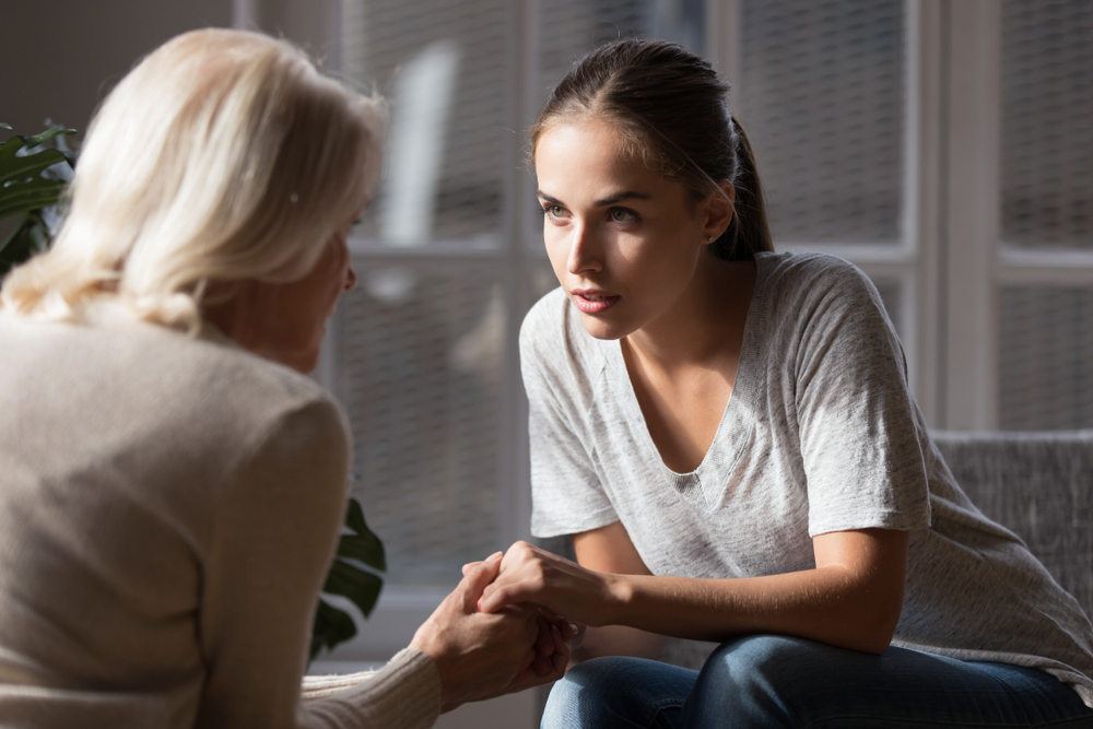 Grown up daughter holds hands of mid aged mother sit on armchair looking at each other having heart-to-heart talk, concept of close relative person, diverse generations connection, mom and adult child