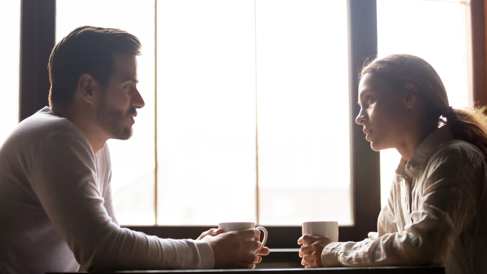 Serious man and woman sitting at cafe table drinking coffee talking and chatting, millennial colleagues or friends spend lunch break in coffeehouse speaking and sharing news enjoying hot beverage