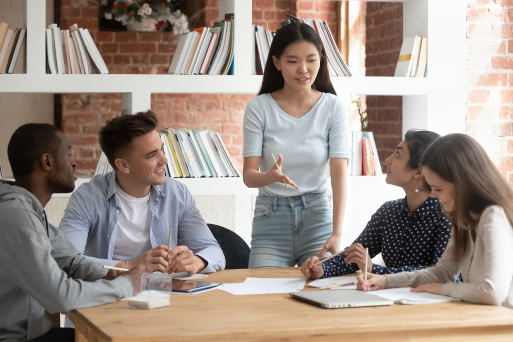 Smiling motivated asian female team leader holding meeting with diverse groupmates, explaining new project detail. African american, indian and caucasian concentrated students discussing assignment.