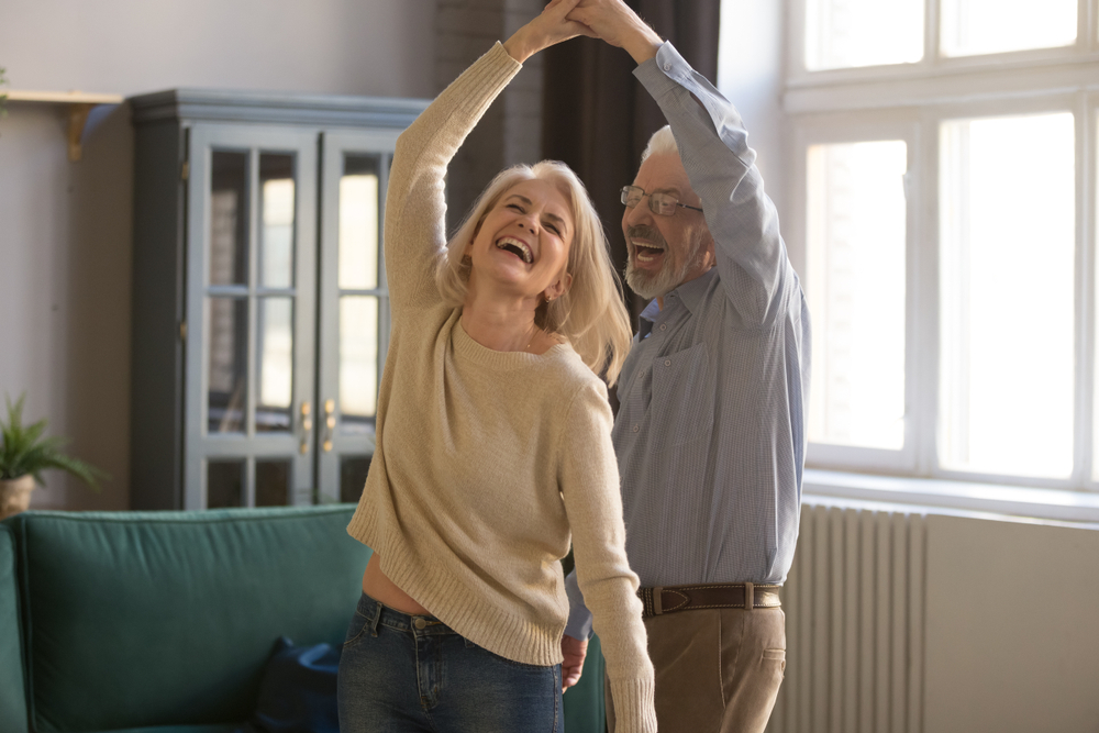 Overjoyed mature grey-haired Caucasian husband and wife have fun enjoy time together at home, happy elderly couple spouses dancing in living room, senior man lead sway smiling middle-aged woman