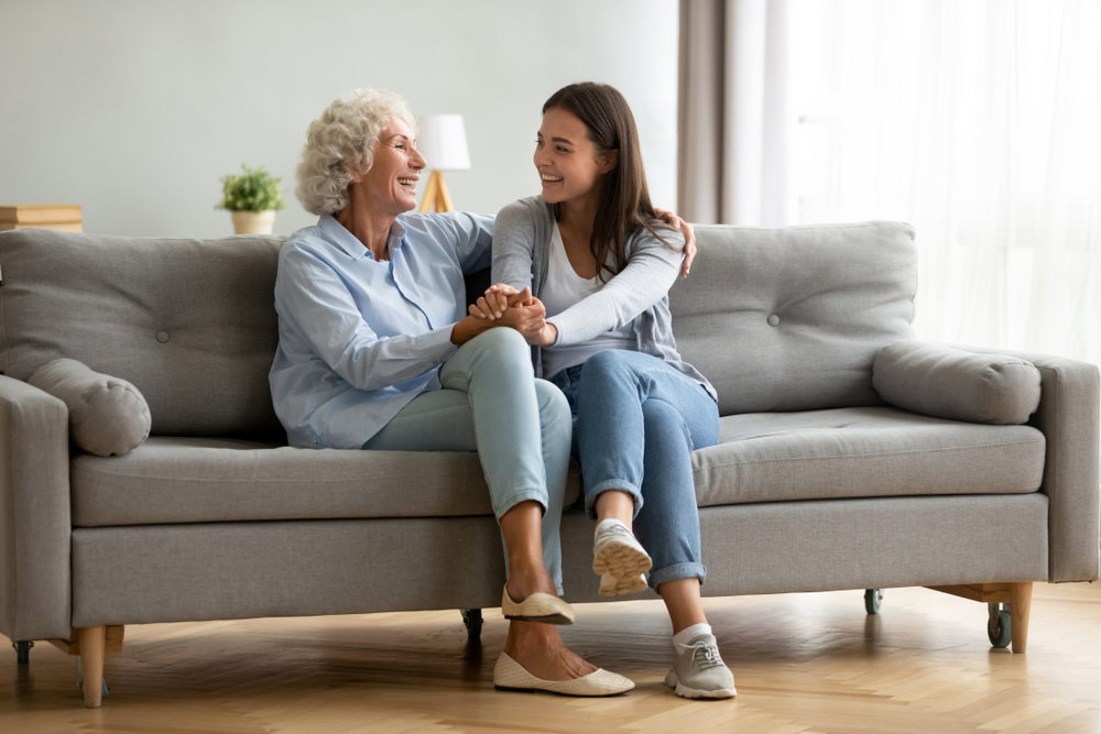 Happy adult granddaughter and senior grandmother having fun enjoying talk sit on sofa in modern living room, smiling old mother hugging young grown daughter bonding chatting relaxing at home together