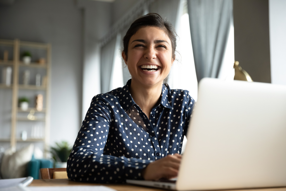 Cheerful indian girl student professional laughing looking away sit with laptop computer at home office table, positive hindu woman having fun enjoy sincere emotion laughter studying working on pc
