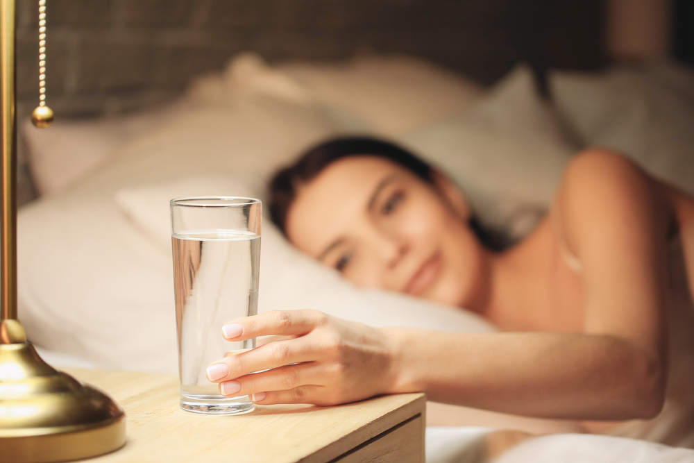 Beautiful young woman taking glass of water from bedside table in evening