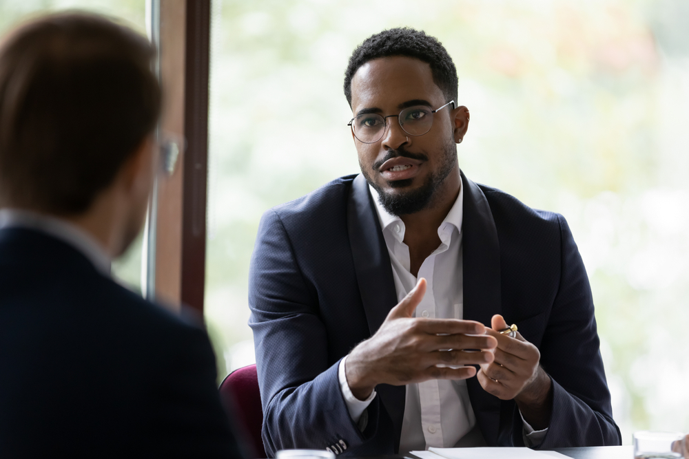 Confident concentrated African American male employee talk with colleague explain thought or idea, focused biracial businessman speak with coworker or partner, brainstorm at office boardroom meeting
