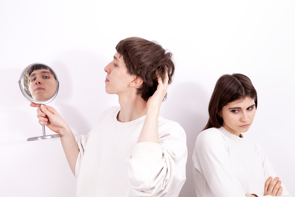 The narcissistic young man in a white sweatshirt is looking at the mirror. He is concentrated on himself and ignoring the young girl standing next to him.