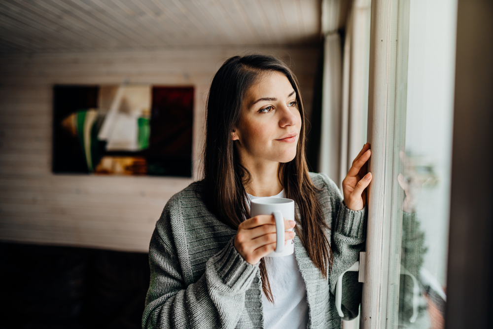 Young woman spending free time home.Self care,staying home.Enjoying view,gazing through to the window.Quarantined person indoors.Serene mornings.Avoiding social contact.