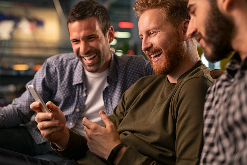 Friends meeting in bar, using smartphone. Happy young friends sitting on couch and watching streaming on mobile phone while laughing. Mid man with friends holding smart phone while watching video.