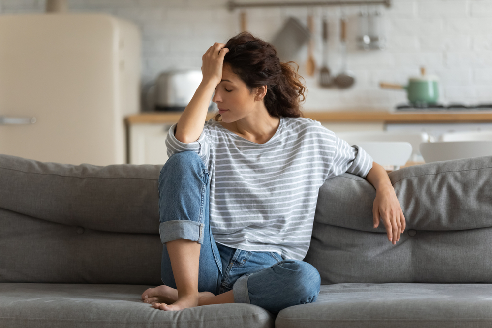 Exhausted frustrated young woman touching forehead, sitting on couch alone, suffering from strong headache or migraine, worried girl thinking about problems, divorce or break up, lost in thoughts
