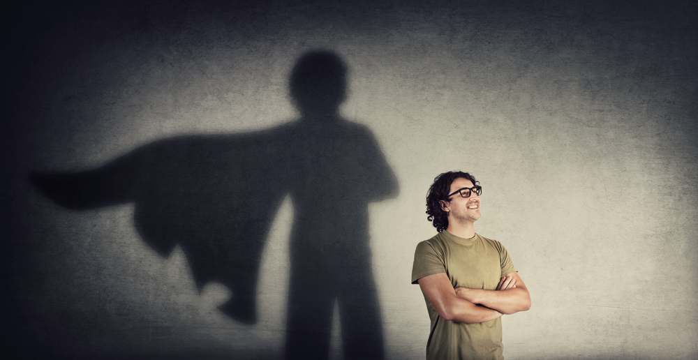 Confident young man keeps arms crossed smiling optimistic as casting a superhero with cape shadow on the wall. Motivated and ambitious guy tends to achieve success. Hero leadership and power concept
