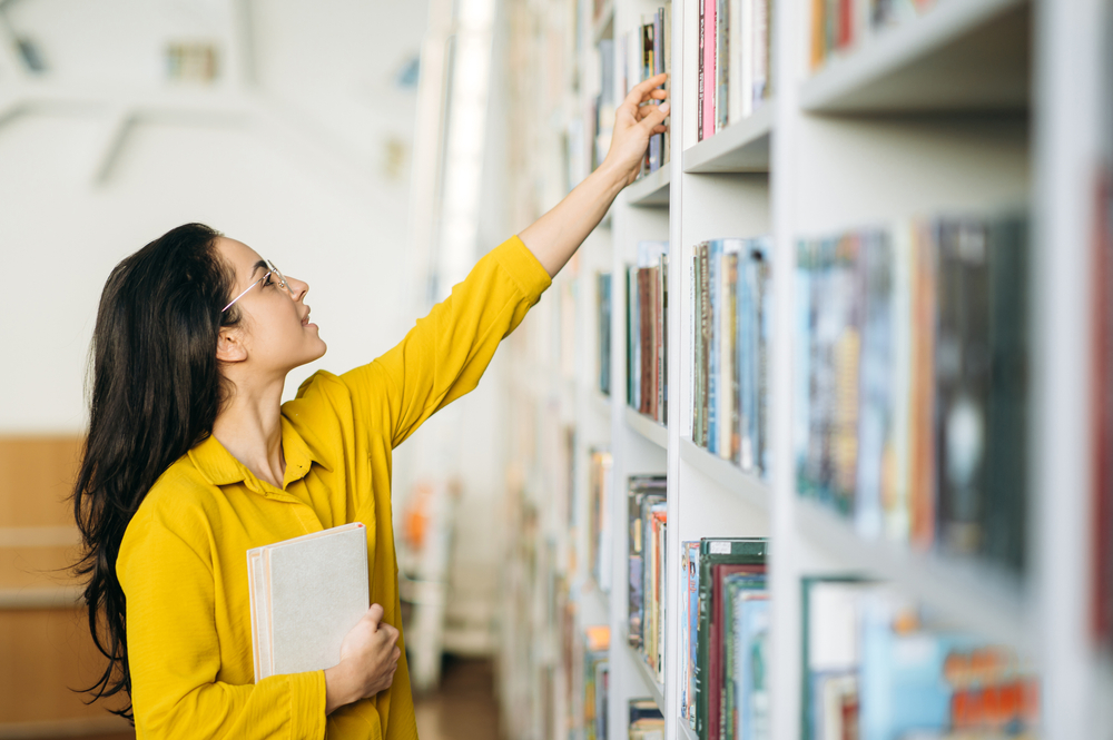 Caucasian female student stands in university library, looking for a book. Beautiful young woman in eyeglasses searching information for a project, learning for passing exams, education concept