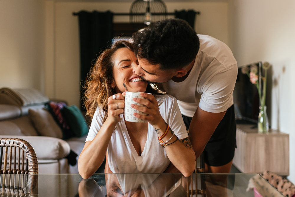 Happy young couple hugging in the living room of their home. Happy couple kissing each other in the face. Family having fun inside a comfortable room having a cup of coffee or tea.