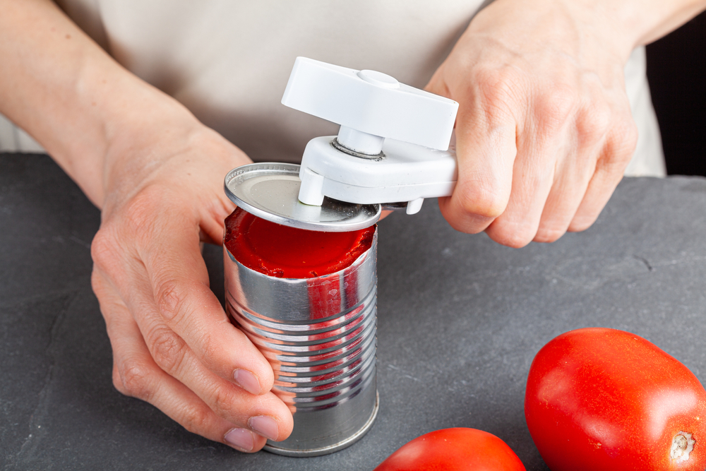 A woman is carefully opening a can of tomato paste on a kitchen counter using a white plastic can opener. She is preparing a meal for which she uses both fresh and pureed preserved tomatoes